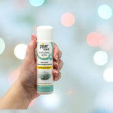A hand holds a bottle of Pjur Med Hydro Glide, a hypoallergenic, water-based personal lubricant. The label highlights "gentle moisturizing." The background features soft, colorful lights in a blur.
